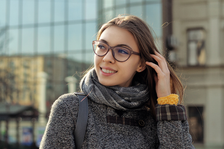 Girl is walking around the city. She has a good mood and she smiles. A student is wearing a coat and a scarf around her neck, the girl is wearing glasses.の写真素材