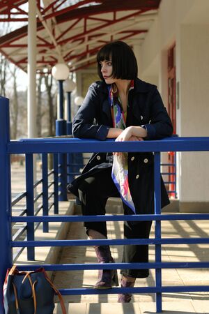 Beautiful girl waiting for transport at the bus stop. She has brown hair and a bob hairstyle, she is wearing a blue raincoat and a scarf around her neck.の写真素材