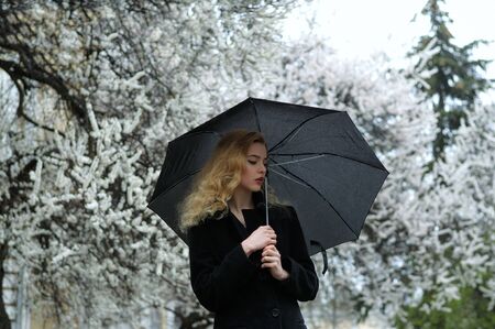 Blonde stands under the umbrella in the rain against the background of flowering apricot treesの写真素材