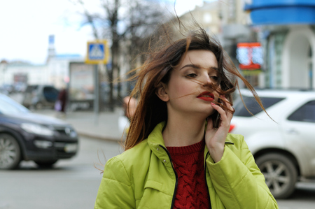 Girl is talking on the phone while walking around the city. She has long brown hair and is dressed in a light green jacketの写真素材