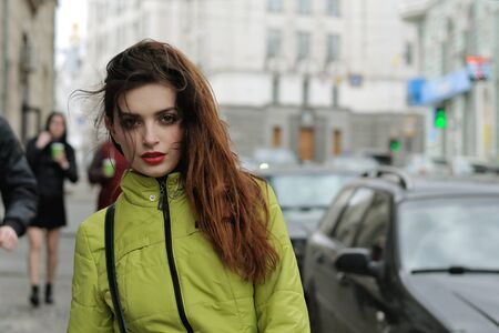Portrait of a stylish girl who walks around the city and the wind blows her long brown hair. She is dressed in a green jacket and has a good mood.の写真素材