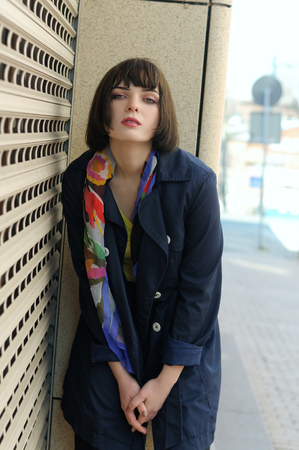 Beautiful girl standing near the closed roller shutters store. She has brown hair and a bob hairstyle, she is wearing a blue raincoat and a scarf around her neck.の写真素材