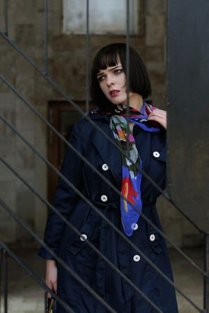 Portrait of a stylish girl near a metal railing. She has brown hair and a bob hairstyle, she is wearing a blue raincoat and a scarf around her neck.の写真素材