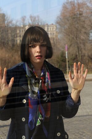 Woman leaned her hands on the window glass. She has brown hair and a bob hairstyle, she is wearing a blue raincoat and a scarf around her neck.の写真素材