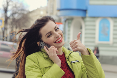 Girl is talking on the phone while walking around the city. She has long brown hair and is dressed in a light green jacketの写真素材