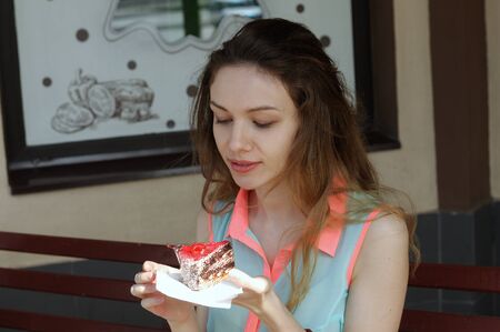 Girl is eating a cake in an outdoor cafe. She holds it with both hands while bringing it to her mouth.の写真素材