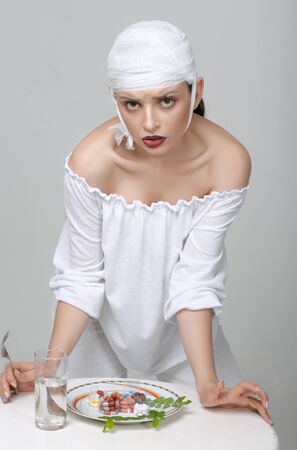 Fashion portrait of a girl with a bandage on her head who stands at the table and has a lunch of medicinesの写真素材