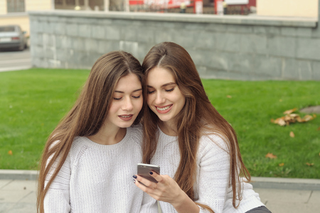 Two brunettes look at photos on their smartphone and laugh. They both have long brown hair that is long to the waist and they are dressed in identical white sweaters.の写真素材