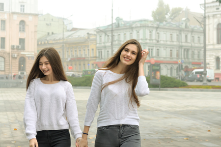 Two girlfriends are walking around the square holding hands and laughing. They both have long brown hair and they are dressed in identical white sweaters against the background of buildings in the fog.の写真素材