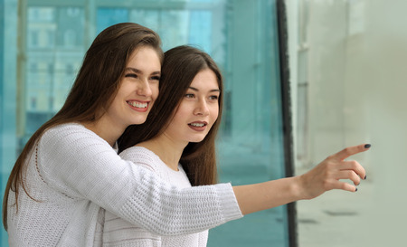 Girl shows her friend a finger at something interesting and smiles. They both have long brown hair that is long to the waist and they are dressed in identical white sweaters.の写真素材