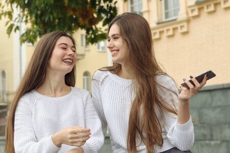 Two brunettes laugh happily during a conversation, waving a smartphone. They both have long brown hair that is long to the waist and they are dressed in identical white sweaters.の写真素材