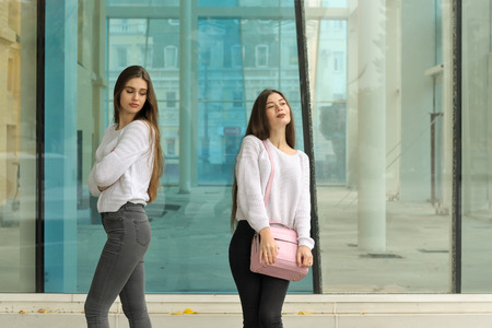 Girl with envy looks at her friend's handbag. They both have long brown hair that is long to the waist and they are dressed in identical white sweaters.の写真素材