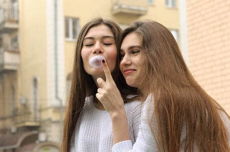 Girl wants to burst bubble of chewing gum. They both have long brown hair that is long to the waist and they are dressed in identical white sweaters.の写真素材