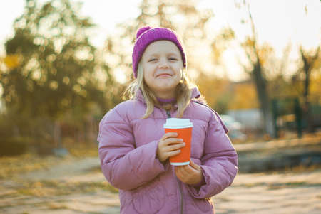 Cute little girl drinks hot chocolate in autumn park in surround of yellowの写真素材