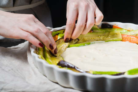 The chef's hands prepare Ratatouille in a baking dish. Side view, black background, close up