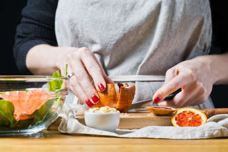 Female chef cuts red oranges for salad with arugula. Black background, side view, kitchen