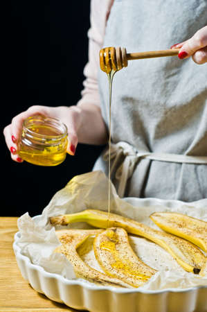 The chef pours honey slices of bananas in a baking dish. Cooking baked bananas. Black background, side view, kitchenの写真素材