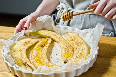 The chef pours honey slices of bananas in a baking dish. Cooking baked bananas. Black background, side view, kitchenの写真素材
