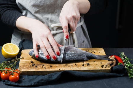 Hands chef cut Dorado fish on a wooden chopping Board, ingredient rosemary, tomatoes, chili, lemon, thyme, pepper, salt. Dark background, side viewの写真素材
