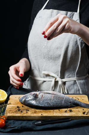 Hands chef salted Dorado fish on a wooden chopping Board, ingredient rosemary, tomatoes, chili, lemon, thyme, pepper, salt. Dark background, side viewの写真素材