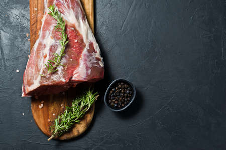 Raw leg of lamb on a wooden chopping Board. Rosemary, thyme, black pepper. Black background, top...