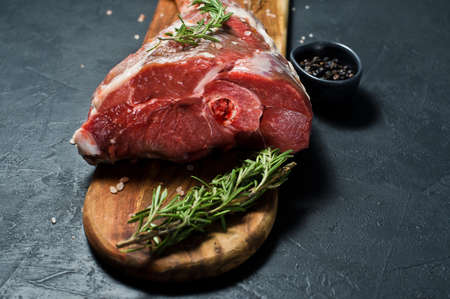 A raw leg of lamb on a wooden chopping Board. Rosemary, thyme, black pepper. Black background, side...