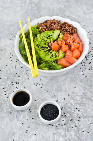 Poke bowl. Ingredients: salmon, avocado, brown rice, algae. Gray background, side view, space for textの写真素材