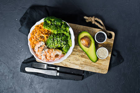 A bowl of balanced health food. Ingredients: shrimps, avacado, carrots, broccoli and rice. Black background, top viewの写真素材