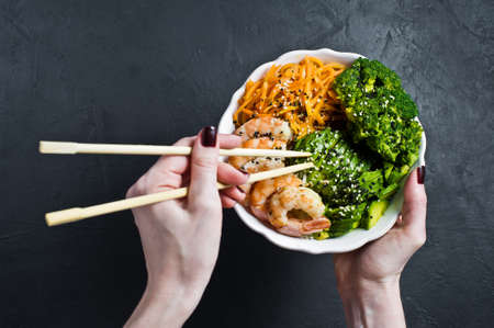 Girl holding Buddha bowl. Ingredients: shrimps, avacado, carrots, broccoli and rice. Black background, top viewの写真素材