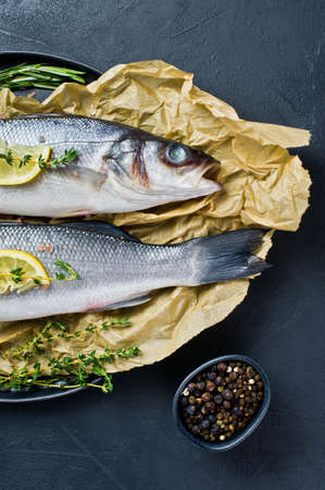 Two raw sea bass on Kraft paper with rosemary, thyme and lemon. Black background, top view