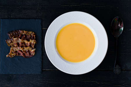 Pumpkin soup with fried bacon in white plate on black background