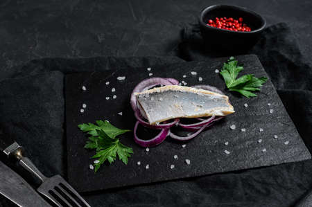 Fillet of salted herring with parsley and red onion. On black rustic background. Top view. Space for...