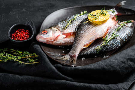 Raw crucian carp with lemon and thyme on a black plate. River organic fish. Black background. Top...