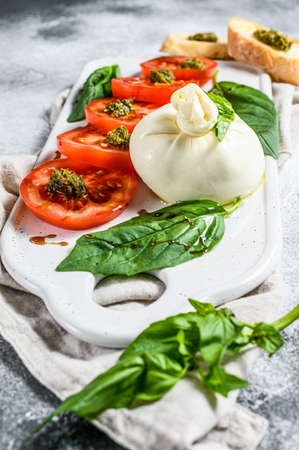 Burrata cheese salad including sliced tomato, basil leaves with pesto. Gray background. Top view.