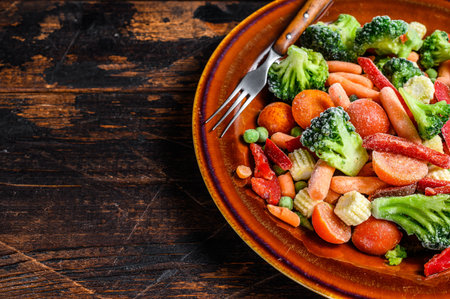 Frozen cut Vegetables, broccoli, sweet peppers, tomatoes, carrots, peas and corn on a plate. Dark Wooden background. top view. Copy space.の写真素材