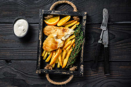 Battered Fish and chips dish with french fries and tartar sauce in a wooden tray. Black wooden background. Top viewの写真素材