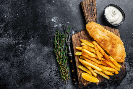 Fish and chips dish with french fries on wooden board. Black background. Top view. Copy space