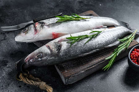 Raw seabass fish on a cutting board with spices, herbs. Black background. Top view