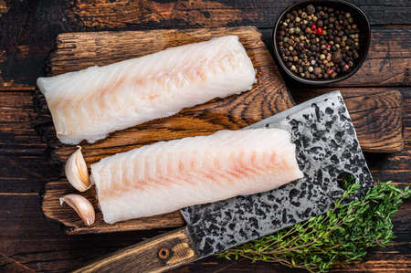 Raw cod loin fillet steak on wooden board with butcher cleaver. Dark wooden background. Top view
