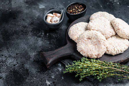 Uncooked Raw breaded patty cutlets on wooden chopping board with herbs. Black background. Top view....