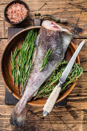 Raw cod or codfish fish in wooden plate with herbs. Wooden background. Top view