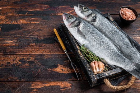 Seabass or sea bass raw fish in a wooden tray with herbs. Dark wooden background. Top view. Copy spaceの写真素材