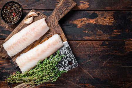 Raw cod loin fillet steak on wooden board with butcher cleaver. Dark wooden background. Top view....