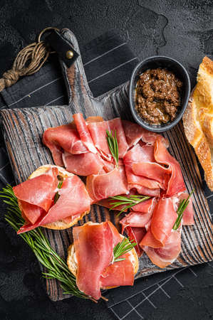 Toast with tomatoes and cured Slices of jamon serrano ham, prosciutto crudo parma on wooden board with rosemary. Black background. Top viewの写真素材