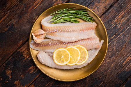 Pollock sea fish fillets, raw whitefish on a plate with herbs and lemon. wooden background. top...