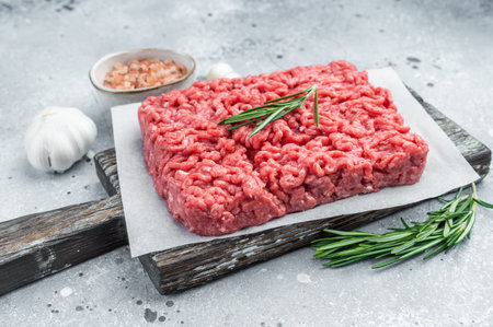 Fresh raw ground mince beef meat with rosemary on parchment over rustic wooden board and concrete...