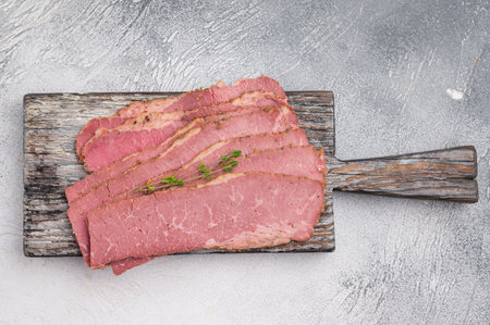 Slices of smoked pastrami placed on an aged wooden cutting board with fresh thyme. Rustic food styling on a textured gray surface, ideal for culinary projects and menusの写真素材