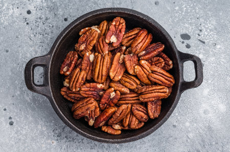 Brown pecan halves displayed in a cast iron pan resting on weathered wood against a grey textured...