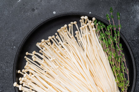 Styled flat lay: Enoki mushrooms, fresh thyme, and garlic on a black plate against a dark, dramatic...