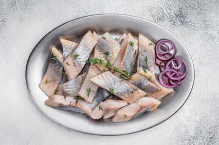 Salted and marinated herring pieces with red onion and thyme served in white dish on wooden board....